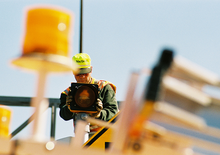 Man wearing MnDOT safety gear holds up a signal lighting component
