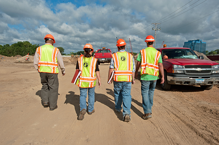 Four engineers wearing safety vests and hardhats walk through a project site with their backs to the camera